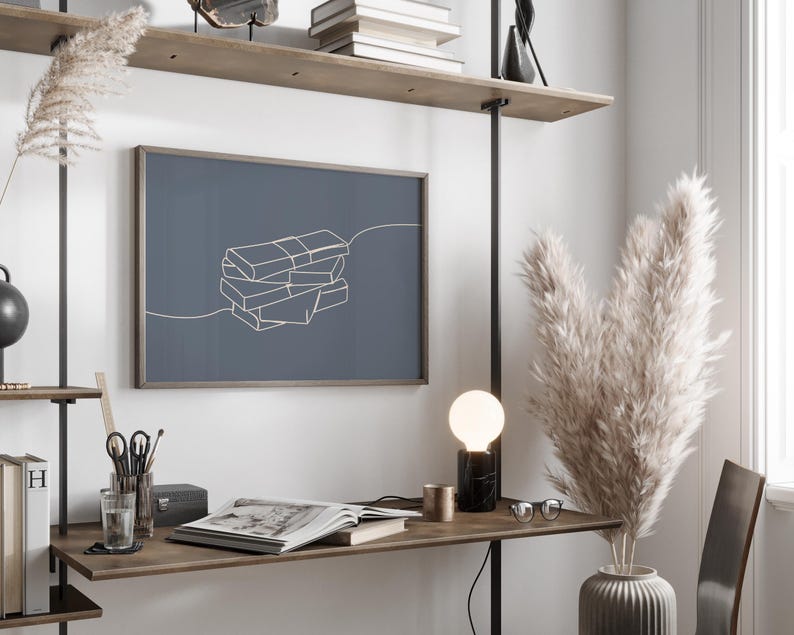 Puede incluir: Una moderna oficina en casa con una impresi&oacute;n enmarcada que presenta un dibujo lineal minimalista de libros apilados. La habitaci&oacute;n incluye un escritorio con una l&aacute;mpara, un libro abierto y objetos decorativos, con plantas altas y secas en un jarr&oacute;n.