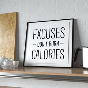 May include: A black framed motivational poster with white text that reads "Excuses - Don't Burn - Calories" on a white background. The poster is sitting on a wooden worktop.