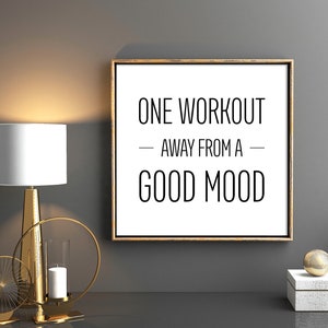 May include: A gold framed motivational poster with the text "ONE WORKOUT - AWAY FROM A - GOOD MOOD" on a white background. The poster is hanging on a gray wall with a gold lamp and decorative candle holders on a black surface.