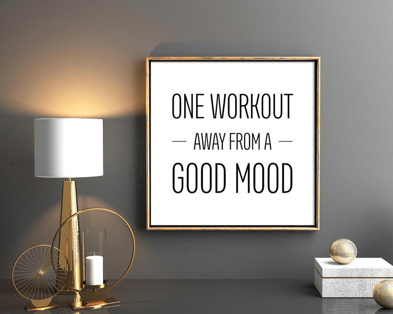 May include: A gold framed motivational poster with the text "ONE WORKOUT - AWAY FROM A - GOOD MOOD" on a white background. The poster is hanging on a gray wall with a gold lamp and decorative candle holders on a black surface.