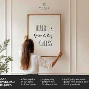 May include: A framed print with the text "HELLO sweet CHEEKS" in black script. The print is held by a person. The frame is a light brown color. The background is a white wall with decorative molding. The Maple Printables logo is in the top right corner.