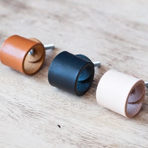 Puede incluir: Tres tiradores de cajón de cuero en marrón, negro y crema. Los tiradores están hechos de cuero enrollado con un tornillo de metal.