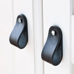 Peut inclure: Deux poignées de tiroir en cuir noir avec une vis argentée au centre de chaque poignée.
