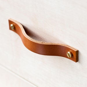 Può includere: Una maniglia per cassetto in pelle marrone con viti in ottone. La maniglia è curva e ha un design semplice.