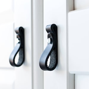 Op de afbeelding: Twee zwarte leren kastgrepen met een eenvoudig ontwerp. De grepen zijn bevestigd aan een witte kastdeur.