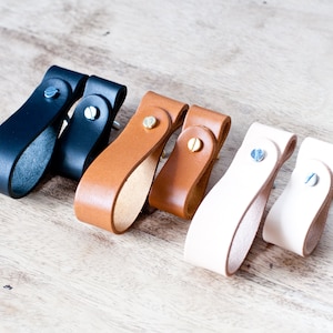 Peut inclure: Cinq poignées de tiroir en cuir de différentes couleurs. Deux sont noires, deux sont brunes et une est beige clair. Chaque poignée a une petite vis en métal au centre.