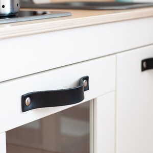 May include: White kitchen cabinet with two black leather handles. The handles have silver metal rivets.