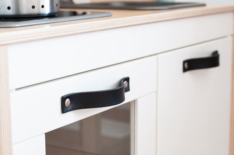 May include: White kitchen cabinet with two black leather handles. The handles have silver metal rivets.