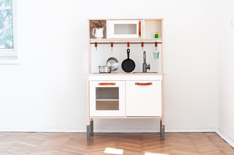 May include: White wooden play kitchen with a black stovetop, a stainless steel sink, and a microwave. The kitchen has a shelf, a cabinet with a glass door, and a cabinet with a solid door. There are hooks for hanging utensils and a small blue bucket.