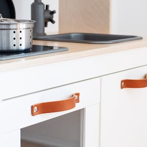 May include: White wooden play kitchen with two brown leather handles. The kitchen has a black stovetop and a stainless steel pot.