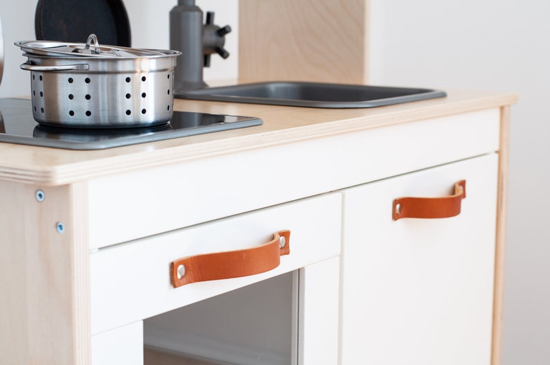 May include: White wooden play kitchen with two brown leather handles. The kitchen has a black stovetop and a stainless steel pot.