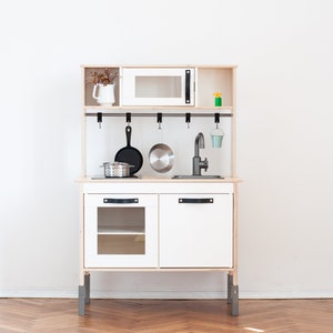Pu&ograve; includere: Una cucina giocattolo in legno con finitura bianca e legno naturale. &Egrave; dotata di forno a microonde, lavello, fornello e pentole e padelle appese. La cucina ha un mobile bianco con maniglie nere e gambe grigie.