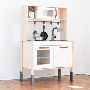 May include: A white wooden play kitchen with a gray sink, a black stovetop, and a white cabinet with a glass door. The kitchen has a wooden shelf with hooks for hanging utensils and a small white cabinet with a glass door. The kitchen is on four gray legs.
