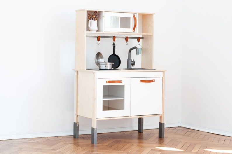 May include: A white wooden play kitchen with a gray sink, a black stovetop, and a white cabinet with a glass door. The kitchen has a wooden shelf with hooks for hanging utensils and a small white cabinet with a glass door. The kitchen is on four gray legs.