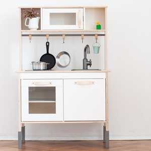 May include: White wooden play kitchen with a black stovetop, a silver sink, and a small oven. The kitchen has a shelf above the oven and a rack with hooks for hanging utensils. There is a small pot, a strainer, and two pans hanging on the hooks.