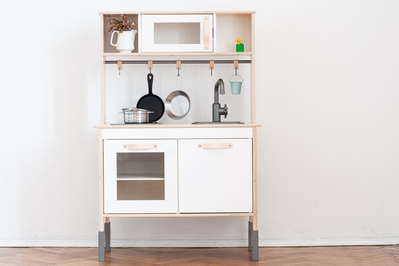 May include: White wooden play kitchen with a black stovetop, a silver sink, and a small oven. The kitchen has a shelf above the oven and a rack with hooks for hanging utensils. There is a small pot, a strainer, and two pans hanging on the hooks.