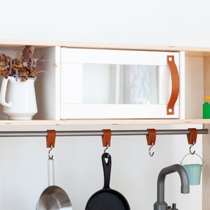 May include: A white play kitchen cabinet with a faux microwave, a metal bar with leather hooks, and a small metal sink. The cabinet has a shelf with a white pitcher and dried flowers.