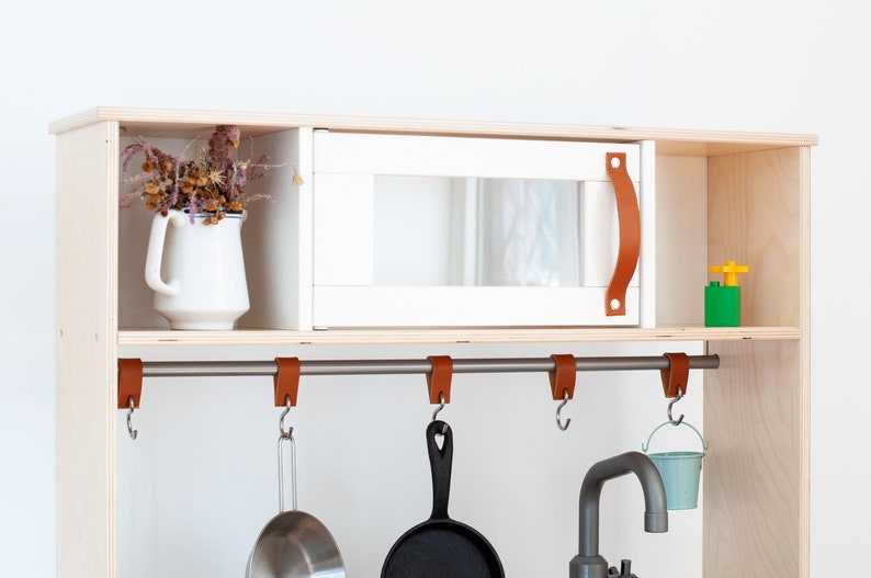 May include: A white play kitchen cabinet with a faux microwave, a metal bar with leather hooks, and a small metal sink. The cabinet has a shelf with a white pitcher and dried flowers.