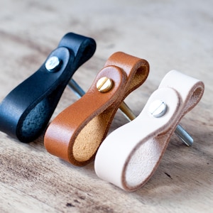 Può includere: Tre tiretti per cassetti in pelle nera, marrone e bianca. Ogni tiretto ha un design a forma di anello e una vite in metallo.