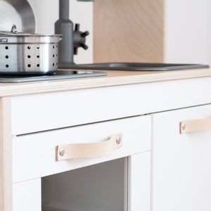May include: White play kitchen with two cabinets, each with a leather handle. The top of the kitchen has a black stovetop and a stainless steel pot with a lid.