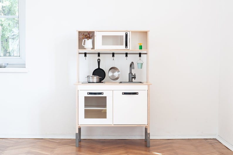 May include: White wooden play kitchen with a microwave, sink, and storage cabinets. The kitchen has a black faucet, a black pot, and a black pan hanging from hooks.