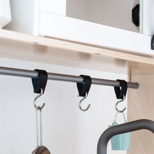 May include: A silver metal bar with black leather straps and hooks hanging from it. The bar is mounted to a wooden shelf. There are three hooks hanging from the bar, with a black pan hanging from one hook and a small blue bucket hanging from another.