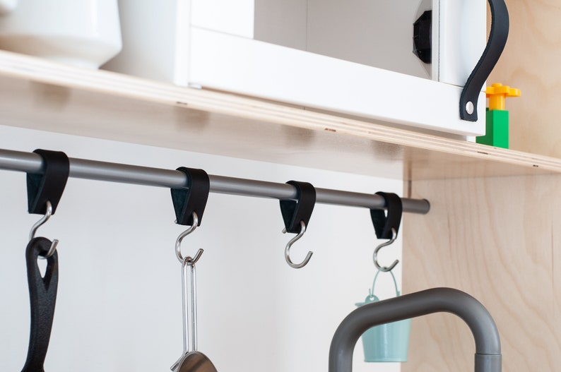 May include: A silver metal bar with black leather straps and hooks hanging from it. The bar is mounted to a wooden shelf. There are three hooks hanging from the bar, with a black pan hanging from one hook and a small blue bucket hanging from another.