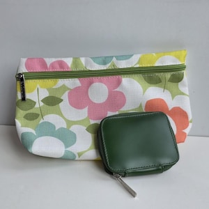 May include: A rectangular fabric pouch featuring a light green background with a pattern of large pink, yellow, orange, and blue flowers, paired with a small, square, dark green zippered case with a silver-toned metal zipper pull.