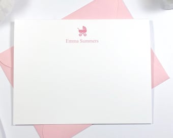 May include: A white card with a pink baby carriage and the text "Emma Summers" printed on it. The card is on a white surface with a pink envelope and a pen.