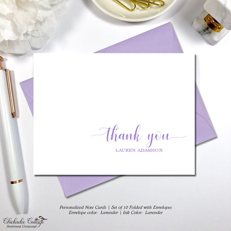 May include: White note card with a lavender "thank you" script and the name "LAUREN ADAMSON". The card is on a lavender envelope. A white pen and a decorative flower are in the background. The text reads "Personalized Note Cards".