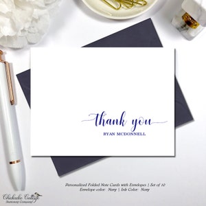 May include: White folded note card with navy blue "thank you" script and the name "RYAN MCDONNELL" in a simple font. The card is on a navy envelope. A white pen and a white flower are in the background.
