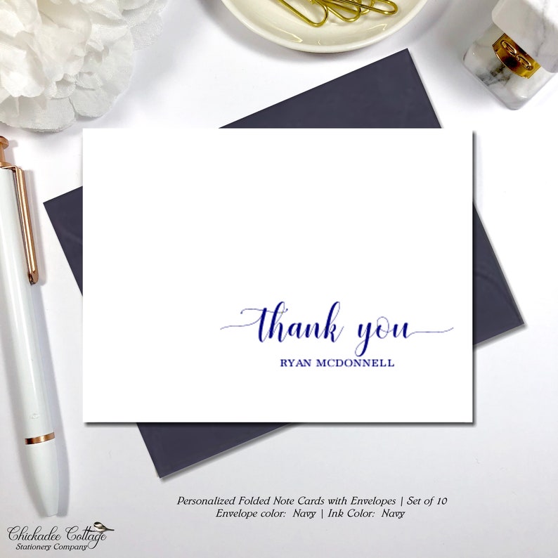 May include: White folded note card with navy blue "thank you" script and the name "RYAN MCDONNELL" in a simple font. The card is on a navy envelope. A white pen and a white flower are in the background.