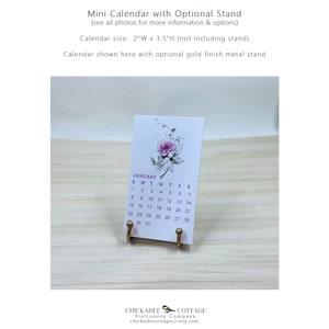 K&ouml;nnte beinhalten: Ein kleiner, wei&szlig;er Kalender mit einem Blumenmuster und dem Monat Januar. Der Kalender steht auf einem goldenen Metallst&auml;nder. Der Kalender ist 5 cm breit und 9 cm hoch.