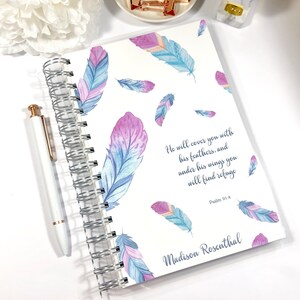 Könnte beinhalten: Ein weißes Spiralblock-Notizbuch mit einer durchsichtigen Plastikhülle. Die Hülle zeigt ein Aquarell-Design mit blauen und violetten Federn und einem Zitat aus Psalm 91,4: "Er wird dich mit seinen Flügeln bedecken, und unter seinen Flügeln wirst du Zuflucht finden." Das Notizbuch ist mit dem Namen "Madison Rosenthal" personalisiert.