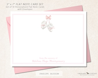 May include: Set of 10 personalized 5x7 inch flat note cards with envelopes. The design features a pair of white baby booties with pink bows hanging from a pink ribbon. The text "from the nursery of Adeline Hope Montgomery" is printed in pink below the booties. The envelope is blossom pink.