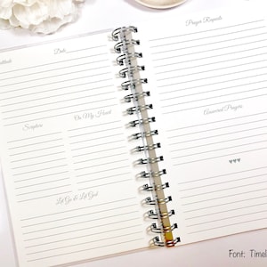 May include: An open spiral bound journal with lined pages for writing. The pages are divided into sections labeled "Gratitude", "Date", "Scripture", "On My Heart", "Let Go & Let God", "Prayer Requests", and "Answered Prayers". The journal has a white background and the text is in gray.
