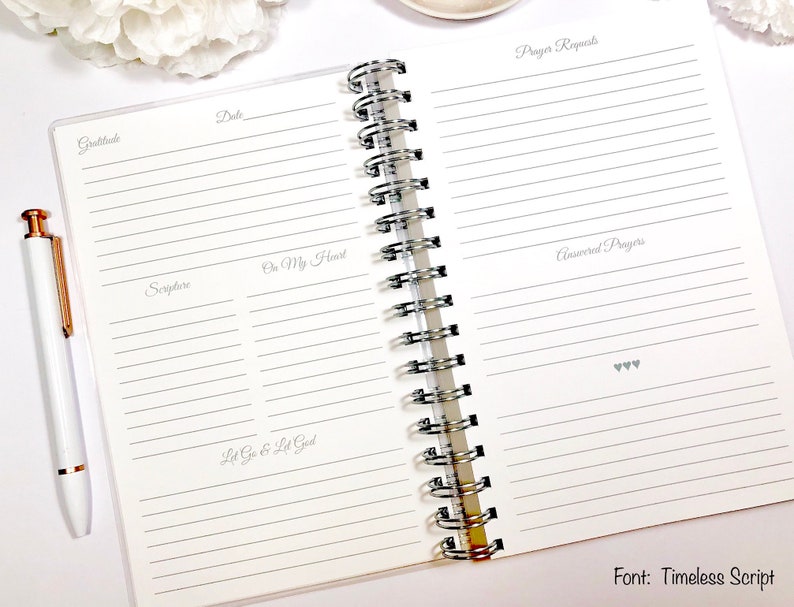 May include: An open spiral bound journal with lined pages for writing. The pages are divided into sections labeled "Gratitude", "Date", "Scripture", "On My Heart", "Let Go & Let God", "Prayer Requests", and "Answered Prayers". The journal has a white background and the text is in gray.
