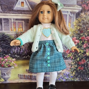 May include: A doll wearing a mint green cardigan, a white shirt with a blue heart, and a teal plaid skirt with matching buttons. The doll has long auburn hair, blue eyes, and a mint green bow. She is wearing white tights and black shoes.