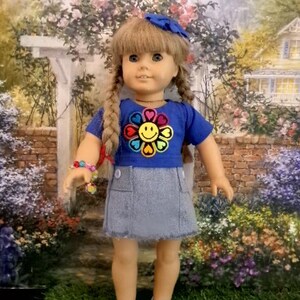 May include: A doll with blonde hair in two braids and a blue hair accessory. The doll wears a blue short-sleeved shirt with a colorful flower and smiley face graphic, a grey denim-style skirt with two front pockets, and light blue clogs.