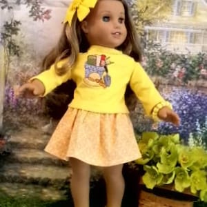 May include: A doll with long brown hair wearing a bright yellow long-sleeve shirt featuring a graphic of a garden basket with tools, a light orange patterned skirt, a yellow hair bow, a yellow beaded bracelet, and yellow perforated clogs.