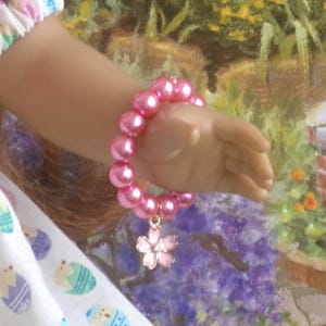 May include: A pink beaded bracelet with a flower charm. The bracelet is made of large, round, pink beads and features a small, pink flower charm. The bracelet is worn on a hand, with a colorful background.