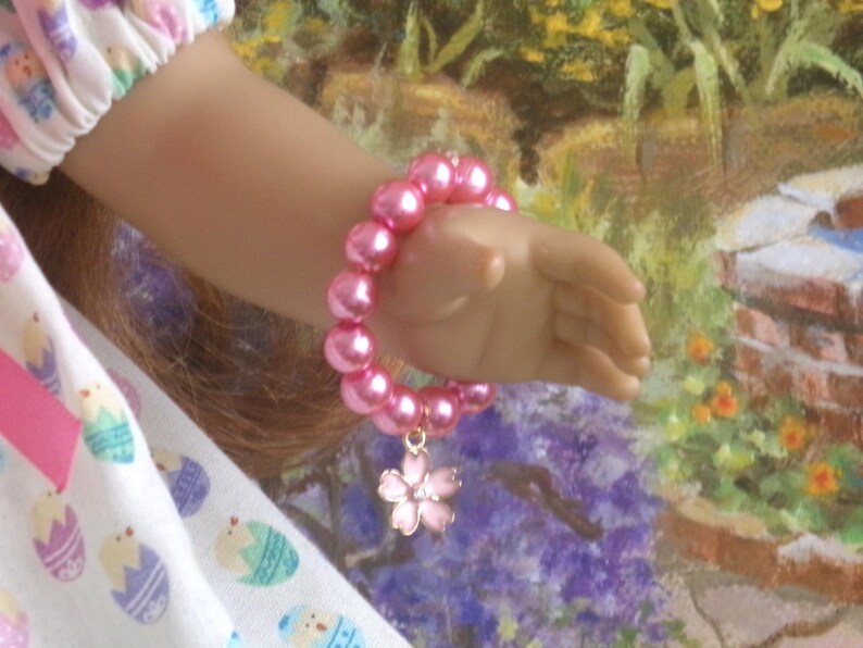 May include: A pink beaded bracelet with a flower charm. The bracelet is made of large, round, pink beads and features a small, pink flower charm. The bracelet is worn on a hand, with a colorful background.