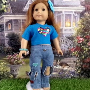 May include: A doll with long brown hair and a blue hair accessory, wearing a bright blue short-sleeved shirt with a multicolored sequin heart graphic on the chest, and blue denim jeans featuring distressed holes, a small teal heart, and a red heart patch.