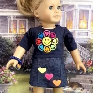 May include: A doll with blonde hair styled in a ponytail, wearing a navy blue short-sleeved shirt featuring a colorful flower and smiley face graphic, a dark denim skirt with three small heart appliques, and navy blue sneakers with white laces.