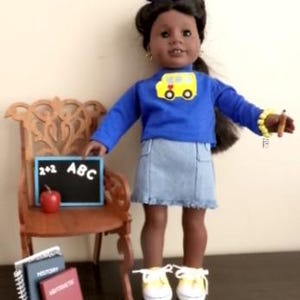 May include: A doll with dark hair styled with a blue bow, wearing a blue long-sleeve shirt with a yellow school bus graphic, a light blue denim skirt, and white sneakers. The doll stands next to a wooden chair with a chalkboard, apple, and books.