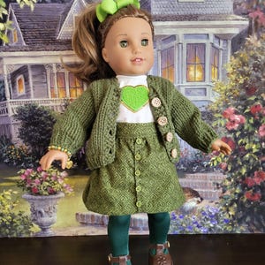 May include: A doll wearing a green outfit, including a sweater, skirt, and tights. The sweater has wooden buttons and a heart-shaped patch. The skirt features a diamond pattern. The doll has brown shoes and a green bow.