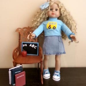 May include: A doll with long curly blonde hair and a light blue bow, wearing a light blue long-sleeve shirt with a yellow school bus graphic, a denim skirt, and blue sneakers. Beside the doll is a brown chair, a chalkboard with 2+2 ABC, a red apple, and books.