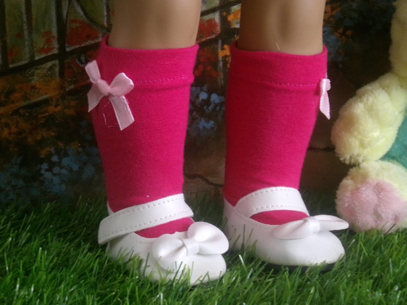 May include: A pair of doll shoes and socks. The shoes are white with a bow on the toe and a strap. The socks are hot pink with a small bow on the side. The items are on a bed of green grass.