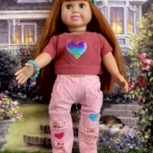 May include: A doll with long auburn hair and a teal hair accessory, wearing a mauve short-sleeved shirt with a holographic heart graphic, light pink distressed pants with heart patches, a teal beaded bracelet, and white lace-up sneakers.