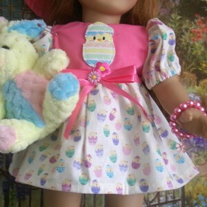 May include: A doll wearing a pink Easter dress with a white skirt patterned with Easter eggs. The doll holds a plush bunny in pastel colors. A pink bow and flower adorn the dress. A pink beaded bracelet is on the doll's arm.